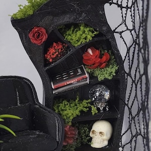 Puede incluir: Una estanter&iacute;a en miniatura negra y curva con varios estantes llenos de musgo verde, flores rojas, dos calaveras blancas, un libro en miniatura titulado I Robot y una peque&ntilde;a bola de cristal. Un sill&oacute;n negro y una red negra est&aacute;n cerca.
