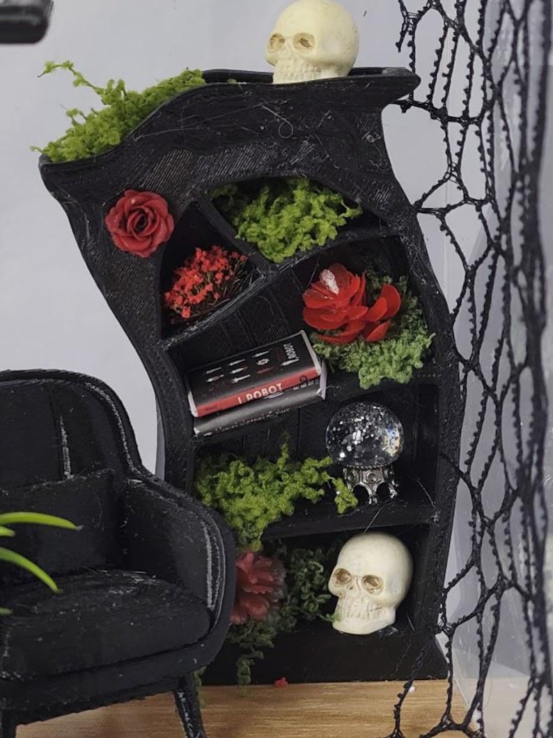 Puede incluir: Una estanter&iacute;a en miniatura negra y curva con varios estantes llenos de musgo verde, flores rojas, dos calaveras blancas, un libro en miniatura titulado I Robot y una peque&ntilde;a bola de cristal. Un sill&oacute;n negro y una red negra est&aacute;n cerca.