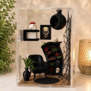 Puede incluir: Una habitaci&oacute;n en miniatura dentro de una caja de acr&iacute;lico transparente con un sill&oacute;n negro, una alfombra redonda negra, una estanter&iacute;a negra con calaveras, rosas rojas y libros, un estante de pared con una calavera, un cuadro y una esfera negra.