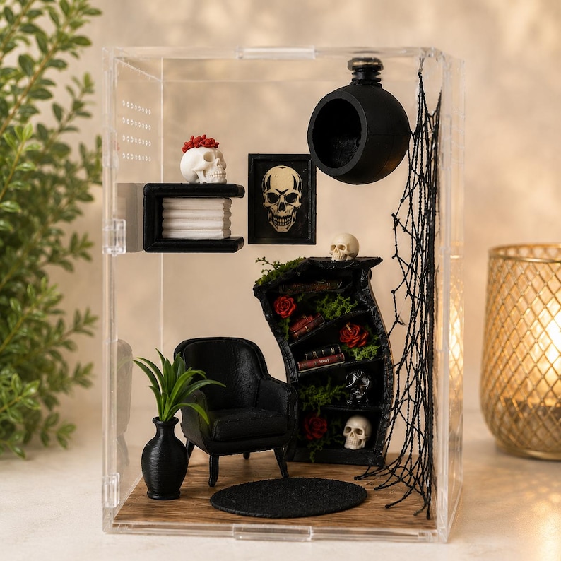 Puede incluir: Una habitaci&oacute;n en miniatura dentro de una caja de acr&iacute;lico transparente con un sill&oacute;n negro, una alfombra redonda negra, una estanter&iacute;a negra con calaveras, rosas rojas y libros, un estante de pared con una calavera, un cuadro y una esfera negra.