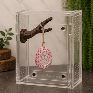 May include: A clear acrylic enclosure featuring a brown textured branch mounted on the interior wall. A light pink, hollow, egg-shaped object with a honeycomb pattern hangs from the branch by a thin cord. The text Tiny Fur Forage is visible.