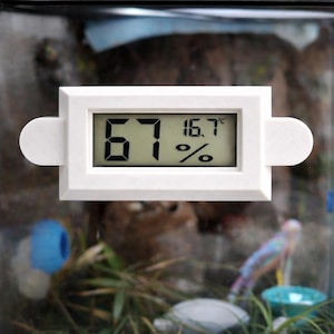 May include: A white rectangular digital hygrometer and thermometer featuring a clear LCD screen displaying the numbers 67 and 16.7 degrees Celsius. The device has rounded side tabs for mounting and is encased in a simple plastic frame.