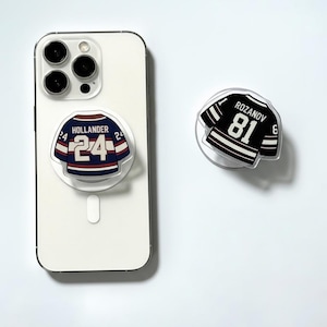 Op de afbeelding: Twee doorzichtige acryl telefoonhouders met hockeytrui-ontwerpen. Eén houder is bevestigd aan een witte telefoon en toont een blauw, wit en rood shirt met "HOLLANDER 24". De andere toont een zwart, wit en rood shirt met "ROZANOV 81".