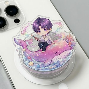 May include: A clear acrylic phone grip with a cartoon figure with purple hair, perched on a pink whale. The whale is decorated with ocean details and rainbow colours. The grip is designed to attach to a phone.