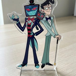 Może przedstawiać: Przezroczysty akrylowy stojak z dwiema postaciami z kreskówek. Jedna postać ma głowę w postaci ekranu telewizora, nosi cylinder i muszkę. Druga postać nosi okulary, garnitur i trzyma laskę. Stojak stoi na drewnianej powierzchni.