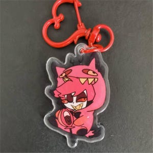May include: A clear acrylic keychain featuring a cartoon character in a pink outfit with a red lobster claw clasp. The character has a pink face, white teeth, and a lollipop. The keychain is a fun accessory.