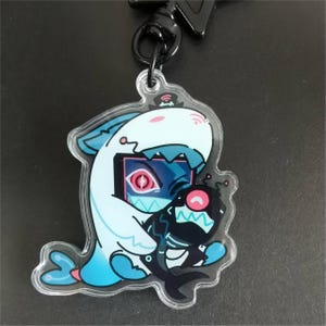 May include: A clear acrylic keychain featuring a cartoon shark wearing a shark costume, hugging a black creature. The shark costume is white and light blue, with a pink eye and a screen-like design. The keychain has a black clasp.