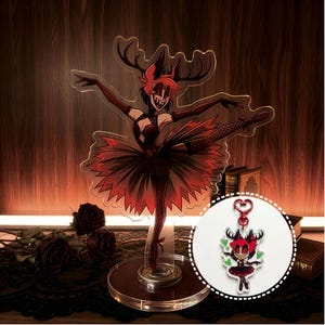 Może przedstawiać: Przezroczysta akrylowa figurka postaci w pozie baletowej, w czerwonej tutu i rogach. Widoczny jest również pasujący brelok z zapięciem w kształcie serca. Figurka stoi na przezroczystym stojaku.