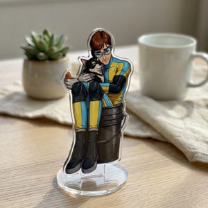 Könnte beinhalten: Klare Acrylfigur einer Person in gelb-blauem Outfit, die einen schwarz-weißen Hund umarmt. Die Figur steht auf einem klaren, kreisförmigen Sockel. Eine kleine Sukkulentenpflanze in einem beigen Topf und eine weiße Tasse sind im Hintergrund.