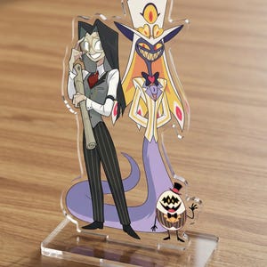 Peut inclure: Support en acrylique représentant des personnages de dessins animés avec une base transparente. Un personnage est une grande silhouette en costume blanc avec un corps violet en forme de serpent. L'autre personnage est une grande silhouette avec de longs cheveux noirs et un gilet gris.