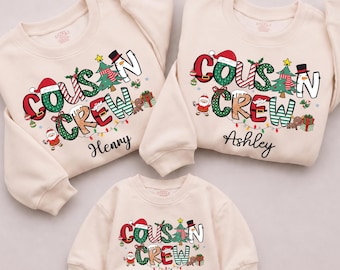 Puede incluir: Tres sudaderas de cuello redondo color crema con el texto "COUSIN CREW" en patrones navideños festivos. Los diseños incluyen gorros de Papá Noel, árboles de Navidad, muñecos de nieve y regalos. Cada sudadera está personalizada con los nombres Henny, Ashley o Sophia.
