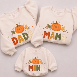 May include: Three cream-colored long-sleeve sweatshirts featuring a graphic of an orange pumpkin with a heart, autumn leaves, and berries. The sweatshirts have the words DADA, MAMA, and MINI printed in large, colorful, stitched-style block letters.