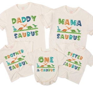 May include: A collection of off-white t-shirts and a onesie with dinosaur-themed graphics. The shirts read "Daddy Saurus", "Mama Saurus", "Brother Saurus", and "Sister Saurus". The onesie says "One A-Saurus".