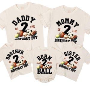 May include: A collection of cream-colored t-shirts and a onesie, each featuring a sports-themed design. The shirts display the words "Daddy," "Mommy," "Brother," and "Sister," along with the number "2" and various sports balls. The onesie says "Born" and "Ball".