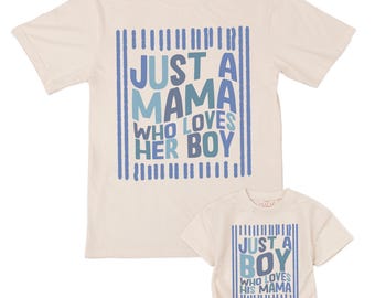 May include: Two matching cream-colored t-shirts. The larger shirt reads "JUST A MAMA WHO LOVES HER BOY" in blue text. The smaller shirt is a onesie with the text "JUST A BOY WHO LOVES HIS MAMA". Both shirts have blue vertical stripes.