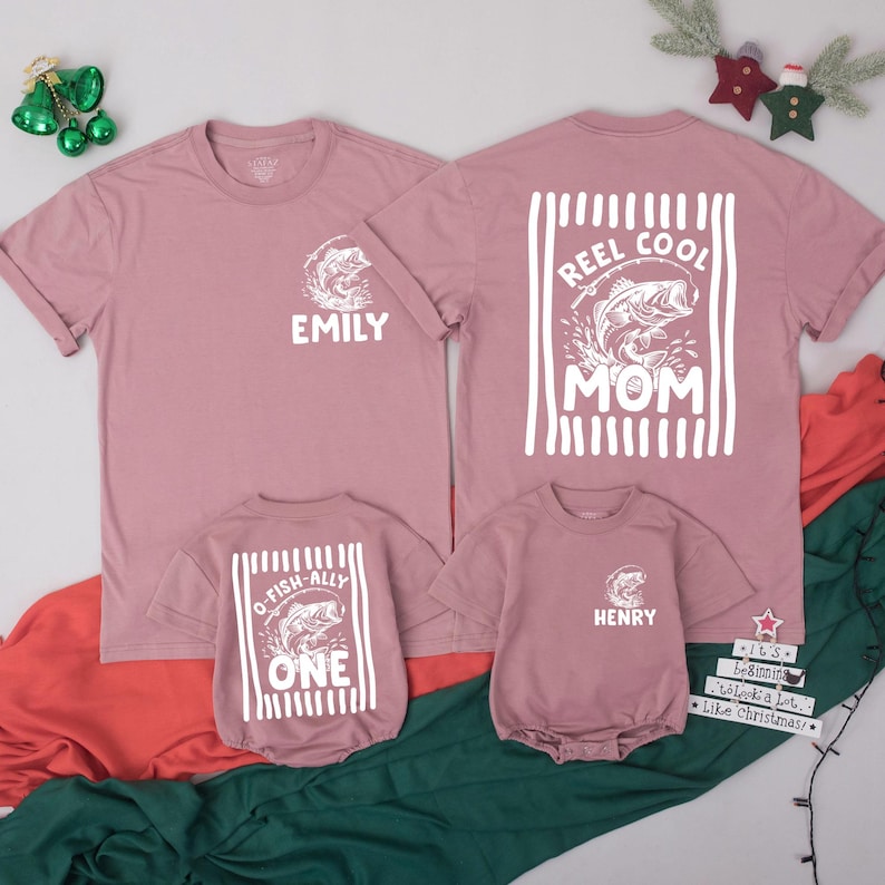 Puede incluir: Dos camisetas malva y dos bodies de beb&eacute; malva con estampados gr&aacute;ficos blancos. Las camisetas presentan una ilustraci&oacute;n de pez y el texto EMILY o REEL COOL MOM. Los bodies presentan una ilustraci&oacute;n de pez y el texto O-FISH-ALLY ONE o HENRY.