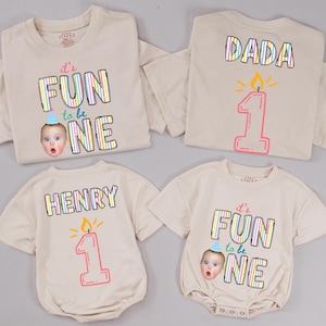May include: Four beige garments including two t-shirts and two baby bodysuits. The designs feature the text "it's FUN to be ONE" with a photo of a baby wearing a party hat, or the text "DADA" or "HENRY" above a pink candle graphic with the number 1.