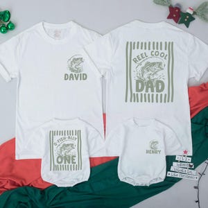 Puede incluir: Dos camisetas blancas y dos bodies de beb&eacute; blancos con gr&aacute;ficos en verde oliva. Una camiseta muestra un pez y el nombre David, la otra un pez y Reel Cool Dad. Los bodies presentan gr&aacute;ficos de peces con el texto O-Fish-Ally One y Henry.
