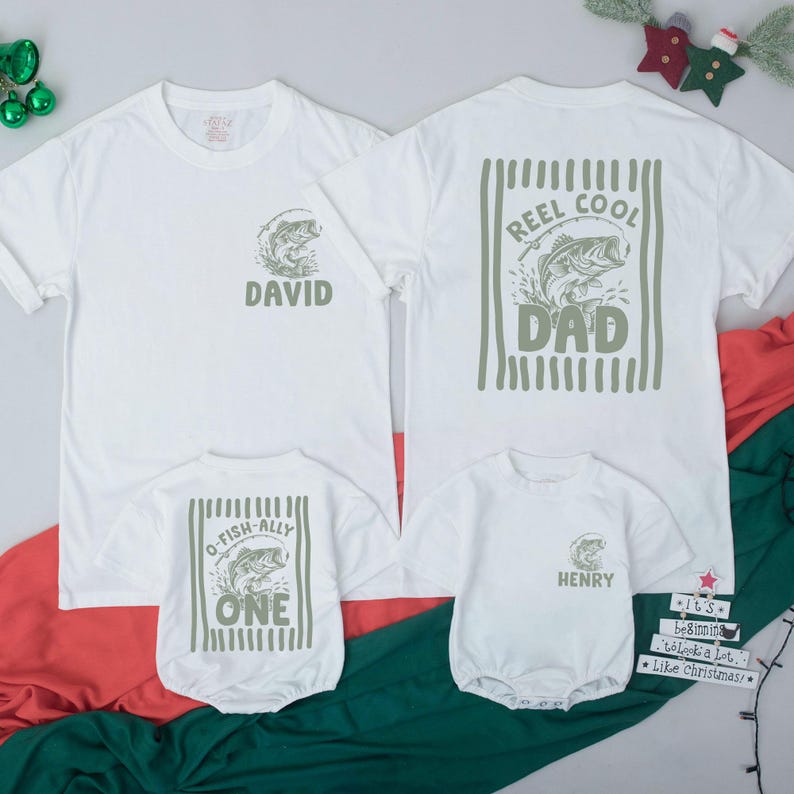 Puede incluir: Dos camisetas blancas y dos bodies de beb&eacute; blancos con gr&aacute;ficos en verde oliva. Una camiseta muestra un pez y el nombre David, la otra un pez y Reel Cool Dad. Los bodies presentan gr&aacute;ficos de peces con el texto O-Fish-Ally One y Henry.