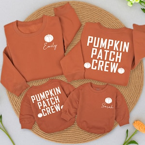 May include: Four rust-colored sweatshirts with white pumpkin graphics. Two sweatshirts say "Pumpkin Patch Crew" and two have a pumpkin and a name. The sweatshirts are arranged on a woven, circular mat, with a few flowers in the background.