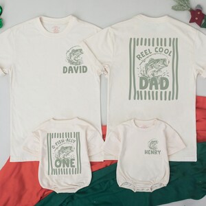 Puede incluir: Dos camisetas y dos bodies de beb&eacute; color blanco hueso con gr&aacute;ficos en verde salvia. Las camisetas de adulto tienen un gr&aacute;fico de pez con el texto DAVID o REEL COOL DAD. Los bodies tienen un gr&aacute;fico de pez con O-FISH-ALLY ONE o HENRY.