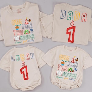 May include: Four cream-colored children's shirts and onesies. Two shirts have the text "ONE FOR THE BOOKS" and "Logan's first birthday". One shirt has "DADA" and the number "1". The onesie has "LOGAN" and the number "1".