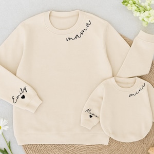 May include: Two cream-colored sweatshirts, one adult and one child size. The adult sweatshirt has "mama" on the chest and "Emily" on the sleeve. The child's sweatshirt says "mini" and "Mama".