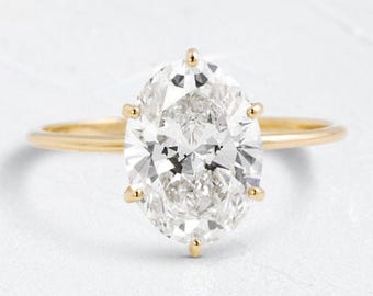 May include: A gold engagement ring with a large, oval-cut diamond. The diamond is set with six prongs and sits atop a thin, gold band. The ring is displayed on a white surface.