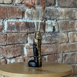 May include: A black and gold ceramic vase in a phallic shape, containing dried flowers. The vase sits on a wooden table against a brick wall. The vase has a glossy finish with gold detailing.