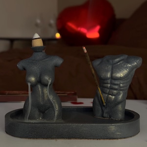 May include: A grey incense holder set with a female and male torso design. The set includes a tray, with an incense stick burning in the male torso. Red heart-shaped decorations are scattered on the surface.