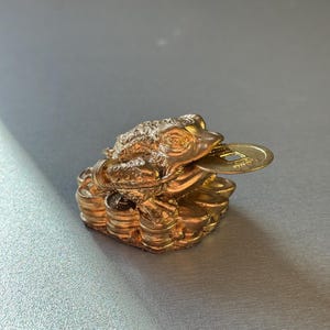 May include: A gold-colored, three-legged toad figurine with a coin in its mouth. The toad sits atop a pile of coins, and the figurine has a textured surface. The background is a solid, light gray color.