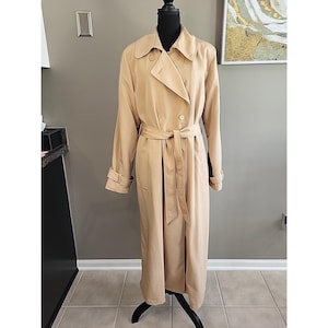May include: A tan trench coat with a double-breasted design, a belt, and button details. The coat is displayed on a black mannequin. The coat has long sleeves and a classic collar.