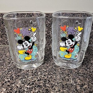 May include: Two clear square-shaped glasses featuring Mickey Mouse pointing upwards. The glasses have a colorful design with Mickey Mouse in the center, surrounded by festive shapes. The year "2000" is printed at the bottom of each glass.
