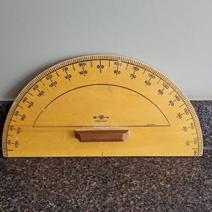 May include: A large yellow semi-circular protractor featuring black degree markings from 0 to 180 in both directions. The tool includes a central wooden handle and the brand name Westcott printed in black text near the center of the arc.