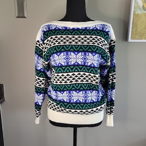 May include: A long-sleeve knit sweater featuring a repeating pattern of white snowflakes, black geometric shapes, and horizontal bands of vibrant blue and green. The garment has a solid white ribbed crew neckline, cuffs, and a matching white hem.
