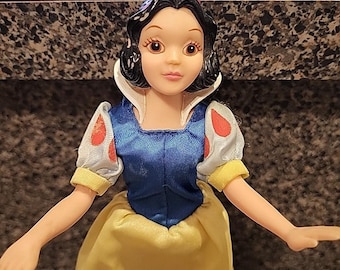 May include: A Snow White doll with black hair, a red bow, and a blue bodice. The doll's sleeves are white with red polka dots, and the skirt is yellow. The doll has a white collar and is a classic Disney character.