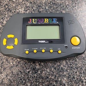 May include: A gray handheld electronic game with the word "JUMBLE" in colorful letters above a screen. The game has yellow buttons and the brand "TIGER" is visible. Buttons include "CLUE", "CLEAR", "SOUND", "OFF", "ON", and "ENTER".