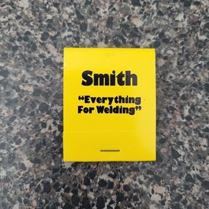 May include: A yellow matchbook with the name "Smith" in bold black letters. Below, the text reads "Everything For Welding" in black. The matchbook is rectangular and has a small black line at the bottom.