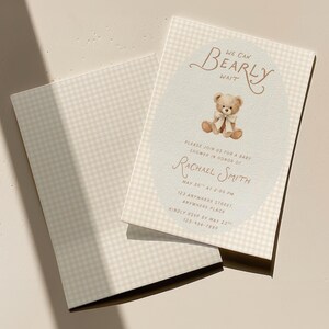 May include: A rectangular baby shower invitation card with a light beige gingham pattern. The center features an oval area with a brown teddy bear illustration wearing a white bow, and text reading We Can Bearly Wait, Please Join Us For A Baby Shower In Honor Of Rachael Smith, May 30th At 2:00 PM, 123 Anywhere Street, Anywhere Place, Kindly RSVP By May 23rd, 123-456-7890.
