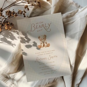 May include: A rectangular baby shower invitation card featuring a light beige gingham pattern, a central illustration of a teddy bear with a cream ribbon, and the text WE CAN BEARLY WAIT, PLEASE JOIN US FOR A BABY SHOWER IN HONOR OF RACHAEL SMITH.