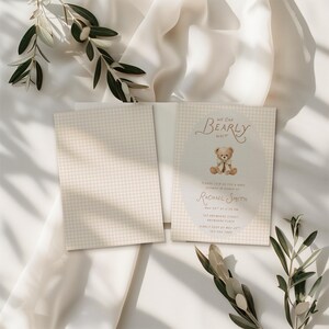 May include: A rectangular baby shower invitation card featuring a beige and white gingham pattern. The card displays a central oval containing a brown teddy bear illustration and the text: We Can Bearly Wait, Please Join Us For A Baby Shower In Honor Of Rachael Smith, May 20th At 2:00 PM, 123 Anywhere Street, Anywhere Place, Kindly RSVP By May 23rd, 123-456-7890.