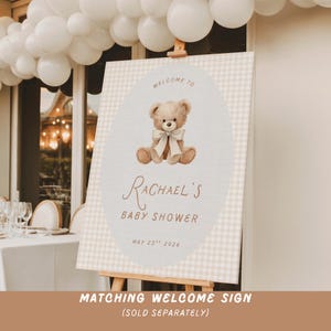 May include: A rectangular welcome sign featuring a beige and white gingham border and a central oval containing a brown teddy bear illustration with a cream bow. Text reads: WELCOME TO RACHAEL'S BABY SHOWER MAY 23RD 2026 in brown lettering.