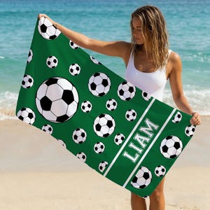 Op de afbeelding: Een rechthoekige strandhanddoek met een donkergroene achtergrond en een herhalend patroon van zwart-witte voetballen. Een witte diagonale streep loopt over het onderste gedeelte, met de naam LIAM in dikke witte hoofdletters.