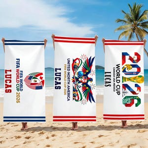 May include: Three white beach towels with FIFA World Cup 2026 designs. Each towel features the name "LUCAS" and different graphics, including flags and the year "2026". The towels are held up on a sandy beach with a palm tree in the background.