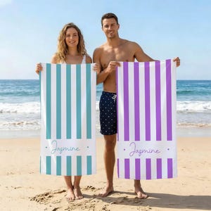 May include: Two beach towels with vertical stripes. One is teal and white, the other is purple and white. Both have the name "Jasmine" printed in cursive. The towels are held by two people on a sandy beach.