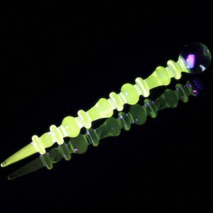 May include: A yellow glass dabber with a round, clear glass tip. The dabber has a series of small, round bumps along its length. The dabber is reflected in a black surface.