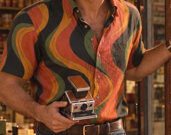 May include: A short-sleeved button-down shirt featuring a wavy pattern in shades of orange, green, and black, paired with dark blue denim jeans, a brown leather belt, and brown leather shoes, while holding a vintage instant camera.