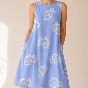 May include: A sleeveless knee-length dress in periwinkle blue featuring a repeating white line-art pattern of stylized fish. The garment has a round neckline, a relaxed A-line silhouette, and side pockets integrated into the side seams.