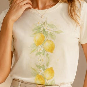 Op de afbeelding: Een cr&egrave;mekleurig T-shirt met korte mouwen en een verticale aquarelillustratie van gele citroenen, groene bladeren en kleine witte bloesems in het midden. Het T-shirt is ingestopt in een lichtbeige broek met een bijpassende riem.