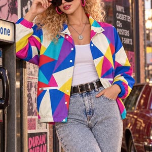 Puede incluir: Una chaqueta cortavientos con un patr&oacute;n geom&eacute;trico vibrante en azul, amarillo, rosa y blanco, sobre una camiseta sin mangas blanca y vaqueros azul claro con efecto lavado &aacute;cido. Incluye gafas de sol negras, aros rosas y collar plateado.
